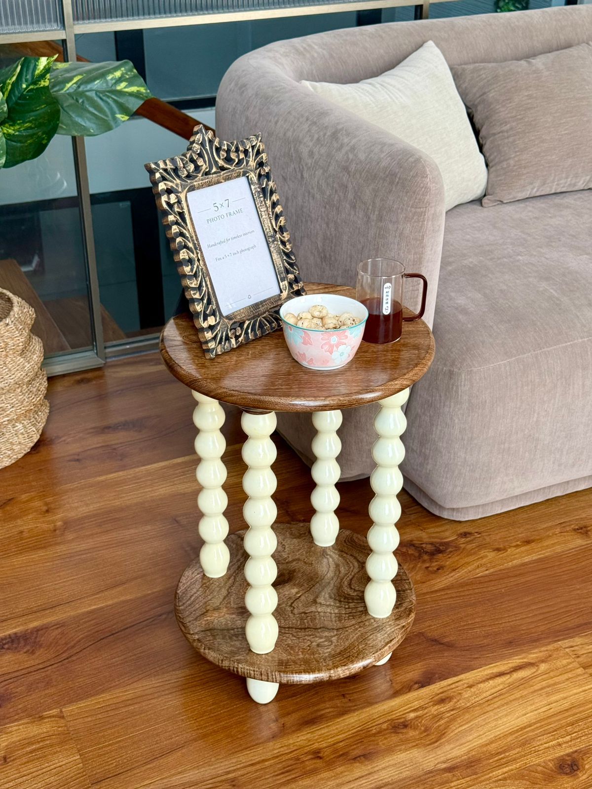 A small round wooden side table with cream-colored turned bead legs, styled in a living room next to a gray sofa with a picture frame and a bowl on top.