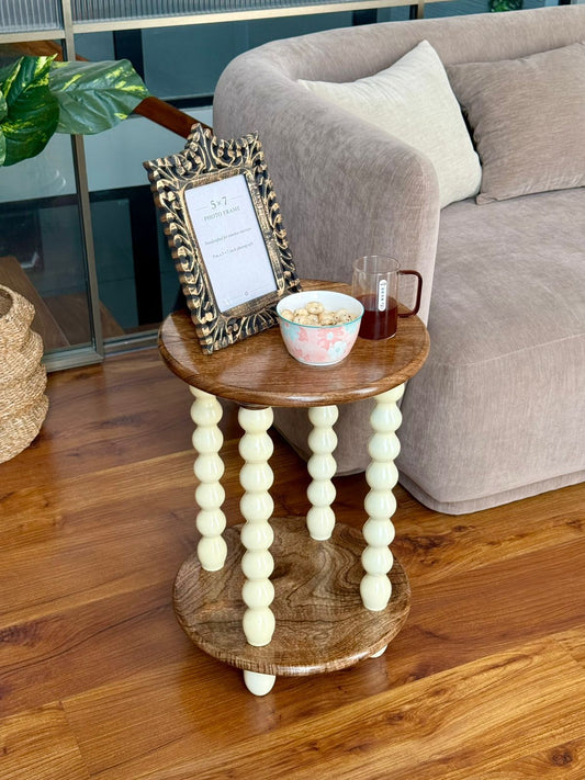 A small round wooden side table with cream-colored turned bead legs, styled in a living room next to a gray sofa with a picture frame and a bowl on top.