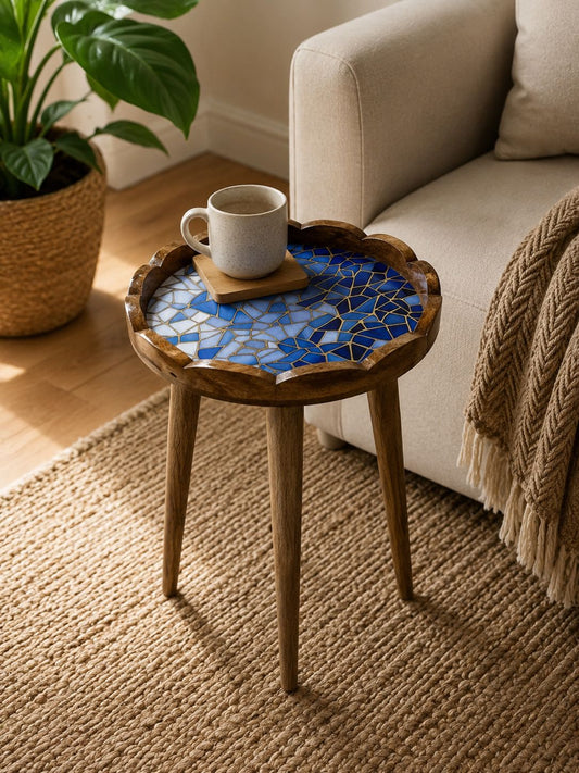 A small round wooden side table with scalloped edges and three legs, featuring a blue mosaic tile top. The table is styled in a cozy living room setting next to a beige sofa with a throw blanket, holding a white ceramic mug on a small wooden coaster.
