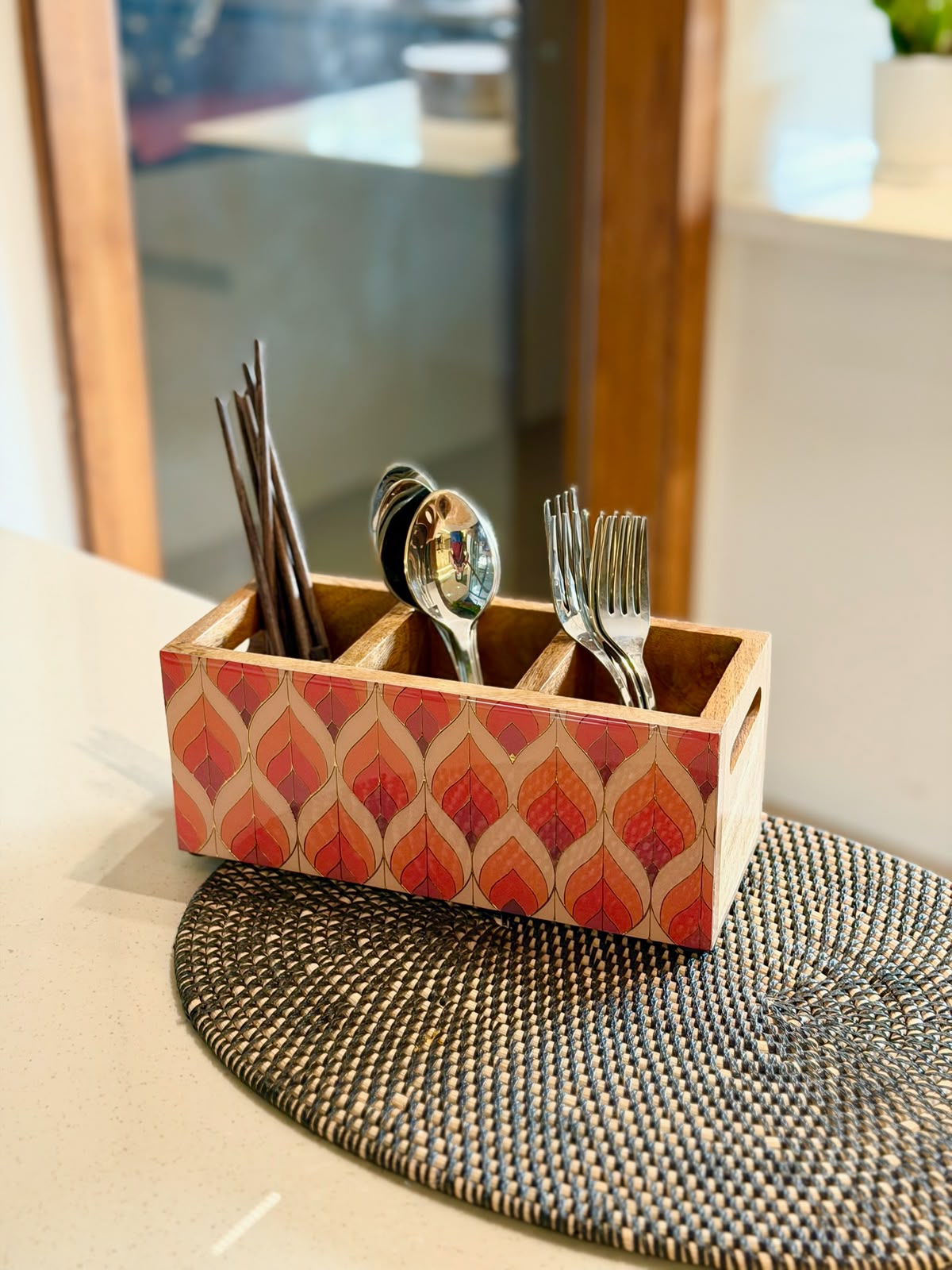 A rectangular wooden utensil holder with a red and beige geometric pattern, containing silver spoons and forks, placed on a woven placemat on a kitchen counter.
