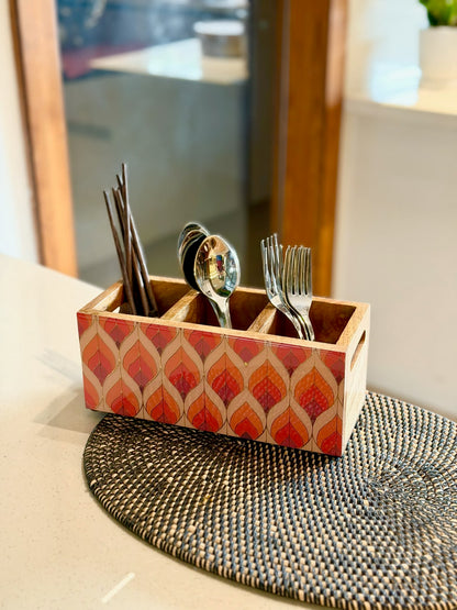 A rectangular wooden utensil holder with a red and beige geometric pattern, containing silver spoons and forks, placed on a woven placemat on a kitchen counter.