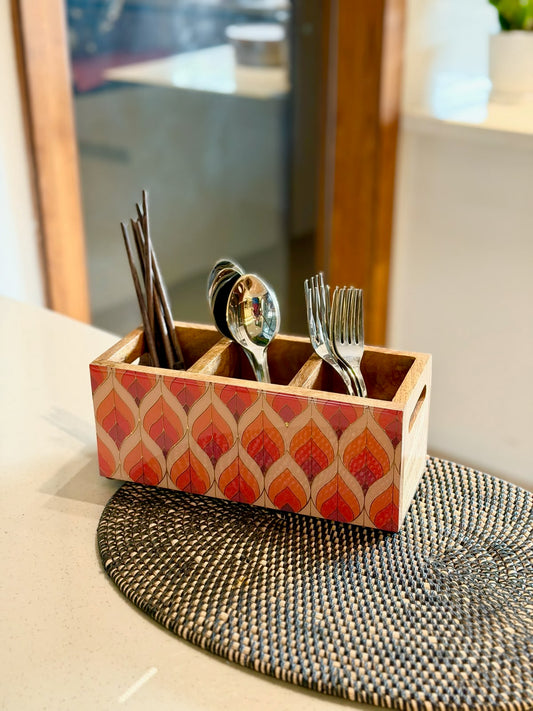 A rectangular wooden utensil holder with a red and beige geometric pattern, containing silver spoons and forks, placed on a woven placemat on a kitchen counter.